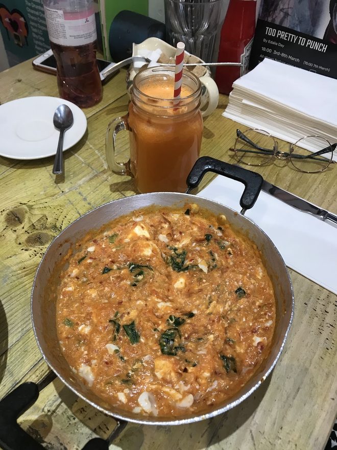 A pan of menemen on a wooden table with an iced coffee
